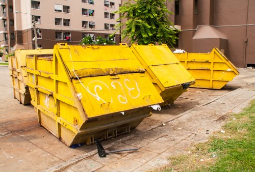 A group of large, yellow metal skip bins with a weathered and slightly dirty finish, positioned on a paved area adjacent to a residential apartment building. The frontmost skip is slightly tilted forward, revealing an open top with visible black support bars and some minor rust spots along its edges. The skip's sides are constructed with sturdy metal panels, and the outer surface displays markings and faded paint, indicative of frequent use. Behind the front skip, there are additional yellow skips aligned in a row, with similar weathered appearances and some slight dents. The background includes a dark brown multi-storey apartment building with balconies and windows, partially obscured by a leafy green tree in the vicinity. To the right, part of a small grassy patch with patches of dirt is visible, showing a typical urban environment suitable for rubbish removal and waste clearance services. The scene is captured in daylight with natural lighting, emphasizing the industrial nature of the waste containers in an urban residential setting, relevant to services provided by Commercial Waste Removal New Cross in the local SE14 postcode area.