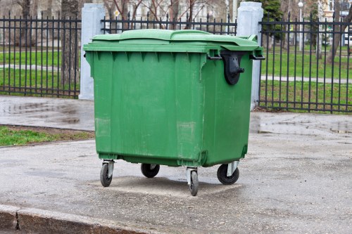 A large, green plastic wheeled rubbish bin with a closed lid positioned on a concrete pavement, with four black caster wheels at the base. The bin has a textured surface with vertical ridges along the sides and a darker, slightly curved lid. The bin is situated outdoors, with a black metal fence and a park with trees and grass visible in the background, indicating a residential or community environment close to New Cross. The area appears wet, possibly after rain, and the surroundings suggest it is used for waste collection in local rubbish removal services, such as those offered by Commercial Waste Removal New Cross.