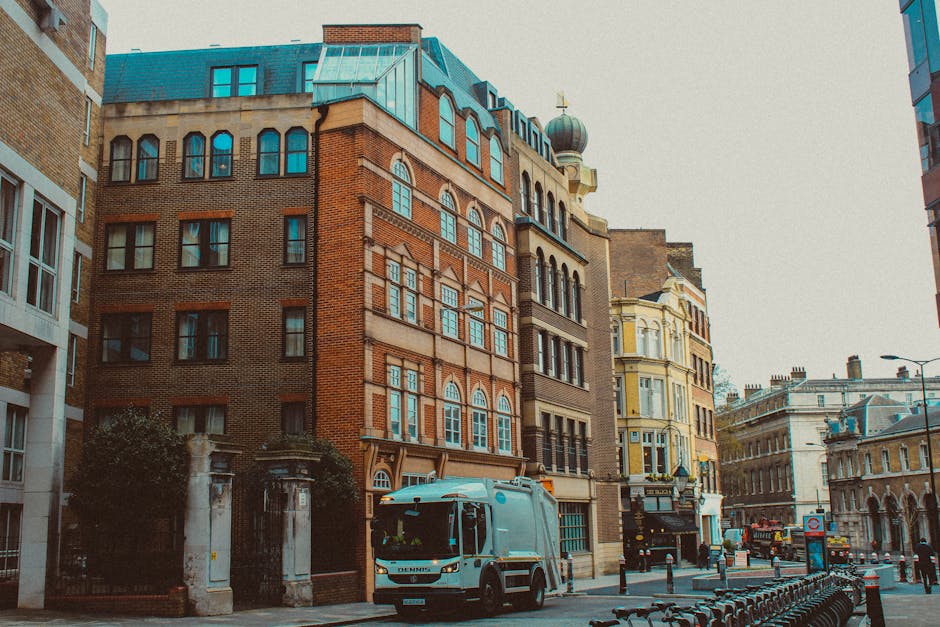 A street scene in an urban area featuring a row of multi-story historic buildings with distinctive architectural details, including arched windows, decorative brickwork, and a domed tower. The buildin