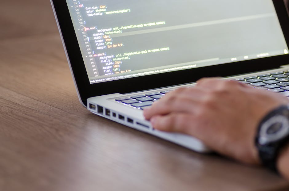 A close-up view of a person's hand resting on a silver laptop keyboard, which is placed on a wooden surface. The laptop screen displays lines of computer code with multicolored syntax highlighting on 